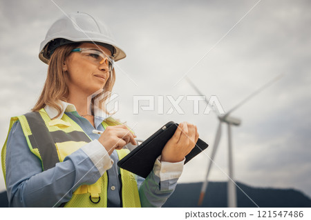 An experienced female engineer is inspecting a wind turbine site, using her tablet to assess the equipments functionality and ensure compliance with strict sustainability standards An experienced female engineer is inspecting a wind turbine site, using her tablet to assess the equipments functionality and ensure compliance with strict sustainability standards 121547486