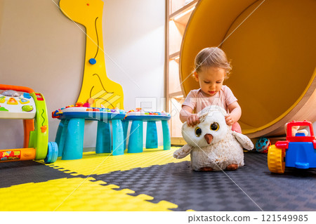 Toddler playing with plush owl toy in colorful indoor playroom with educational activities and toys 121549985