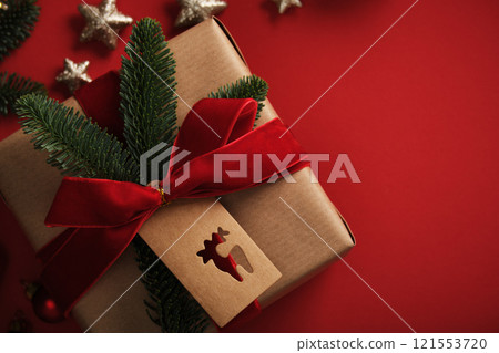 Christmas gift wrapped in kraft paper with red velvet ribbon, a spruce branch and a tag with a silhouette of a reindeer on a red background. Christmas gift wrapped in kraft paper with red velvet ribbon, a spruce branch and a tag with a silhouette of a reindeer on a red background. 121553720