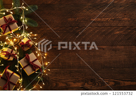 Christmas gifts in brown kraft paper with red velvet ribbons among sprigs of evergreen fir trees and garlands on brown wooden background. Place for your text. Christmas gifts in brown kraft paper with red velvet ribbons among sprigs of evergreen fir trees and garlands on brown wooden background. Place for your text. 121553732