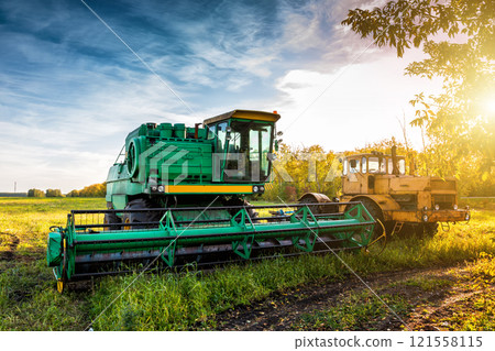 The modern green combine harvester and yellow heavy duty tractor on agricultural field in the rays of the autumn sun 121558115