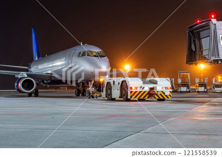 Tow tractor pushes passenger aircraft from boarding bridge at night airport apron 121558539