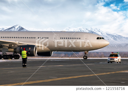 Airport marshaller meets wide body passenger airplane on the background of high snow covered mountains 121558716