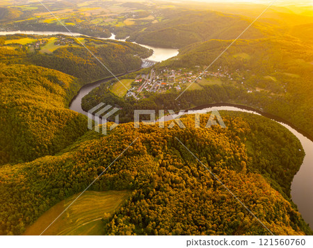 The Slapy Water Reservoir nestled in lush greenery reflects the sunset glow, showcasing the surrounding hills and winding Vltava River, highlighting the beauty of Czechia's natural landscape. 121560760