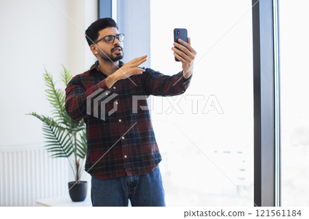 Indian young man holding smartphone near large window, gesturing while taking selfie or video call. Casual setting with natural light and indoor plant creating relaxed ambiance. 121561184