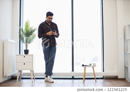 Young Indian man in casual attire using smartphone in modern office environment near large window with natural light. 121561225