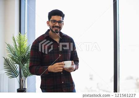 Young Indian man in casual attire smiling while holding coffee in bright office setting. Indoor plant and large window create relaxed, friendly atmosphere. Glasses add touch of modern style. 121561227