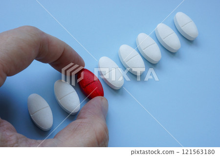 Hand picking a red pill from a row of ordinary ones. Choice and individuality. 121563180