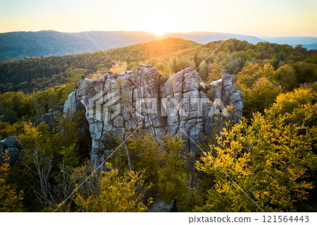 Aerial view of sun sets behind rugged rock formation, casting warm glow over landscape. Trees in autumn colors surround rocks, creating vivid contrast. Dovbush Rocks, Carpathian mountains, Ukraine. Aerial view of sun sets behind rugged rock formation, casting warm glow over landscape. Trees in autumn colors surround rocks, creating vivid contrast. Dovbush Rocks, Carpathian mountains, Ukraine. 121564443