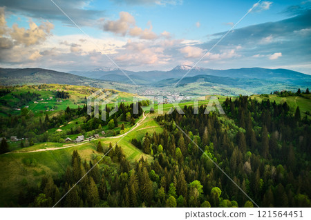 Aerial view of picturesque rural landscape in Ukraine, with lush green fields, dense forests, and scattered houses under sky filled with dramatic clouds. Snow-capped mountains in distant horizon. Aerial view of picturesque rural landscape in Ukraine, with lush green fields, dense forests, and scattered houses under sky filled with dramatic clouds. Snow-capped mountains in distant horizon. 121564451