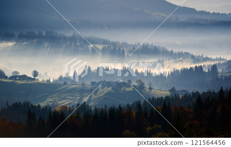 Morning sunlight streams through mist, illuminating serene mountain village nestled among forested hills. Light creates ethereal rays that gently touch autumn foliage and houses scattered on slopes. 121564456