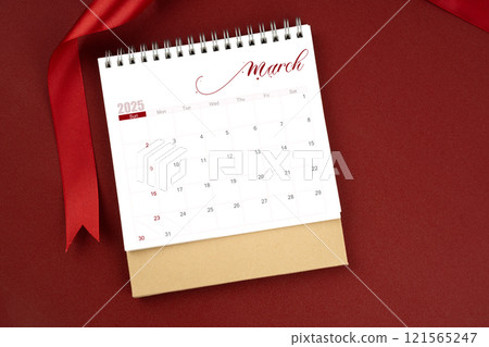 March 2025 desk calendar and red ribbon on red background. 121565247