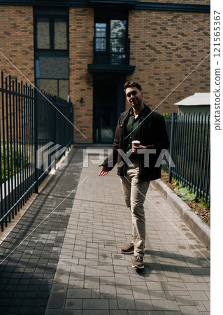 Full length vertical portrait of handsome young man holding coffee cup standing by residential building with metal fence and brick walls, smiling looking at camera, enjoying peaceful moment outdoors. Full length vertical portrait of handsome young man holding coffee cup standing by residential building with metal fence and brick walls, smiling looking at camera, enjoying peaceful moment outdoors. 121565367