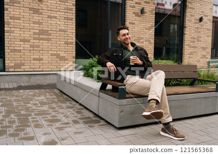 Cheerful young man sitting on city bench outside by brick building, savoring cup of coffee while enjoying pleasant atmosphere of urban environment on sunny autumn day. Concept of urban lifestyle. Cheerful young man sitting on city bench outside by brick building, savoring cup of coffee while enjoying pleasant atmosphere of urban environment on sunny autumn day. Concept of urban lifestyle. 121565368
