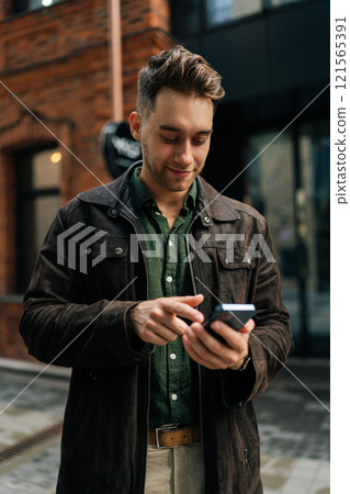 Vertical portrait of happy handsome young man wearing stylish suede jacket using smartphone, smiling looking to screen while enjoying summer day on city street lined with beautiful red brick buildings Vertical portrait of happy handsome young man wearing stylish suede jacket using smartphone, smiling looking to screen while enjoying summer day on city street lined with beautiful red brick buildings 121565391