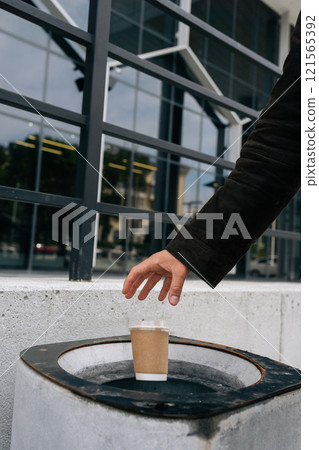 Close-up vertical cropped shot of unrecognizable busines man throwing disposable coffee cup into street trash can near office building, demonstrating responsible waste disposal and urban cleanliness. Close-up vertical cropped shot of unrecognizable busines man throwing disposable coffee cup into street trash can near office building, demonstrating responsible waste disposal and urban cleanliness. 121565392
