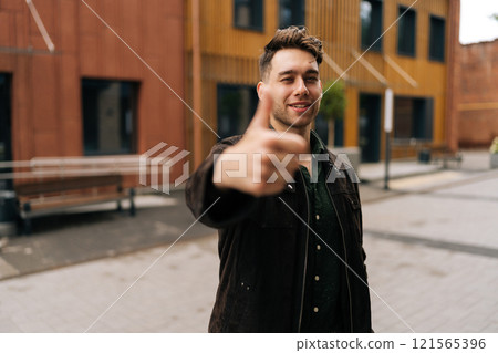 Confident young man wearing casual clothes showing thumbs up gesture of approval, agreement, like or recommendation, standing outdoors in urban street, looking at camera with confidence expression Confident young man wearing casual clothes showing thumbs up gesture of approval, agreement, like or recommendation, standing outdoors in urban street, looking at camera with confidence expression 121565396