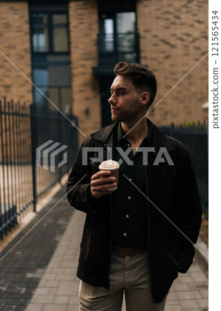 Vertical portrait of confident man pensive looking away while holding coffee cup, enjoying drink standing in urban alleyway by brick buildings, embodying casual city lifestyle and morning tranquility. Vertical portrait of confident man pensive looking away while holding coffee cup, enjoying drink standing in urban alleyway by brick buildings, embodying casual city lifestyle and morning tranquility. 121565434