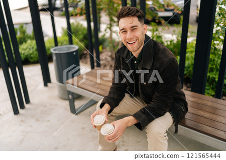 Cheerful handsome young man in casual attire enjoying peaceful moment on park bench in city, holding two coffee cups and laughing looking at camera, surrounded by green trees on beautiful summer day. 121565444