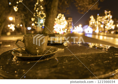 Close-up of a cup of coffee and a slice of cake on a wet table at night. Festive golden bokeh lights create a warm atmosphere. Ideal for holiday, winter, or cozy night-themed projects. Close-up of a cup of coffee and a slice of cake on a wet table at night. Festive golden bokeh lights create a warm atmosphere. Ideal for holiday, winter, or cozy night-themed projects. 121566034