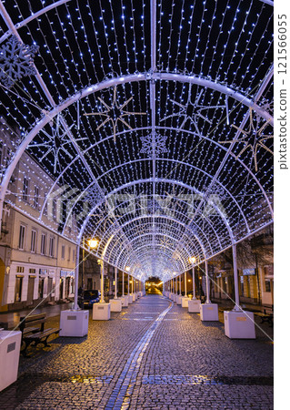 A magical winter scene of a city street adorned with twinkling Christmas lights, creating a tunnel of light. People are walking through the festive atmosphere. 121566055