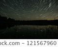 Star trails illuminate the night sky over a calm lake surrounded by a forest during a serene evening 121567960