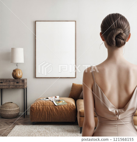 A young woman stands with her back turned to us, gazing at an empty frame on the wall of a modern beige room 121568155