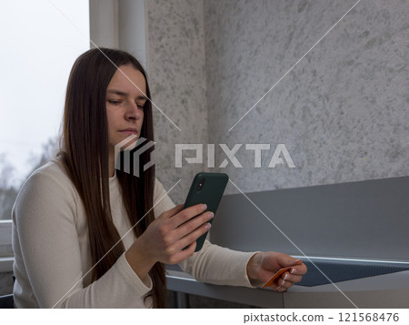 Young woman focusing on smartphone screen, gripping credit card, completing online banking transaction in cozy home environment 121568476