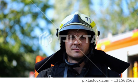 Portrait of confident male fireguard with dirty face standing near a fire engine. Young fireman in full equipment posing after fire against the background of big red truck. Concept of saving lives Portrait of confident male fireguard with dirty face standing near a fire engine. Young fireman in full equipment posing after fire against the background of big red truck. Concept of saving lives 121568562