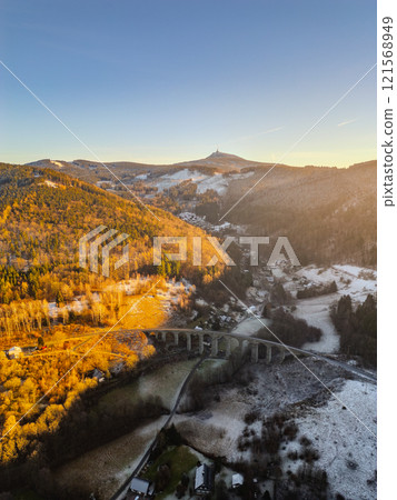 A railway stone bridge arches over a snow-covered landscape in Novina, with the peaks of Jested Mountain glowing in the warm light of a winter sunset. Nature showcases its beauty. A railway stone bridge arches over a snow-covered landscape in Novina, with the peaks of Jested Mountain glowing in the warm light of a winter sunset. Nature showcases its beauty. 121568949