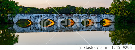 A stone inundation bridge from the late 18th century reflects in the calm waters of Vitek Pond in South Bohemia during a serene evening, surrounded by lush greenery. 121569296