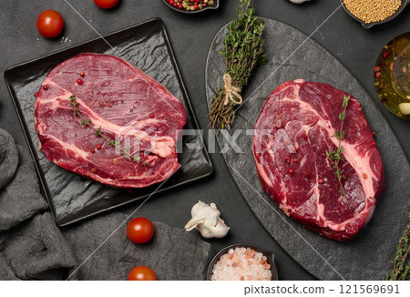 A wooden board holds a raw chuck eye roll steak, complemented by Himalayan salt, thyme sprigs, and peppercorns 121569691