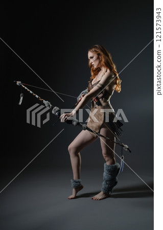 Woman in traditional attire aims an arrow with a bow in a dimly lit studio Woman in traditional attire aims an arrow with a bow in a dimly lit studio 121573943