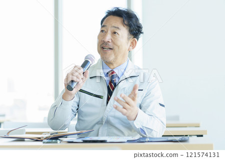 A middle-aged man in work clothes speaking into a microphone. Photo courtesy of Tokyo Electronics College, Denpa Gakuen Corporation. 121574131