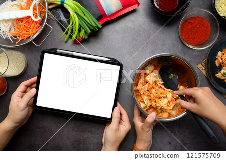 Man holding digital tablet while woman mixing kimchi in stainless steel bowl on a dark countertop 121575709