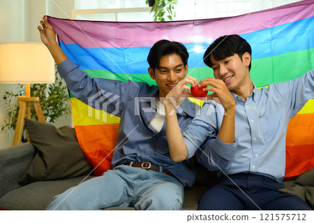 Cheerful young men couple holding a red heart and sitting under rainbow pride flag. 121575712