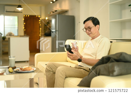 Smiling senior man using a credit card for online payment with mobile phone at home 121575722