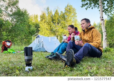 Couple sits on grass near tent in lush forest, enjoying meals from packaged pouches. Dressed in outdoor gear, with portable camping stove and backpack nearby. Green trees and foliage on background. 121577547