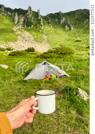 Hand holds white enamel mug filled with hot beverage in front of tent pitched on lush green meadow. Backdrop of steep, rocky cliffs covered in vibrant greenery under cloudy sky. Hand holds white enamel mug filled with hot beverage in front of tent pitched on lush green meadow. Backdrop of steep, rocky cliffs covered in vibrant greenery under cloudy sky. 121577551