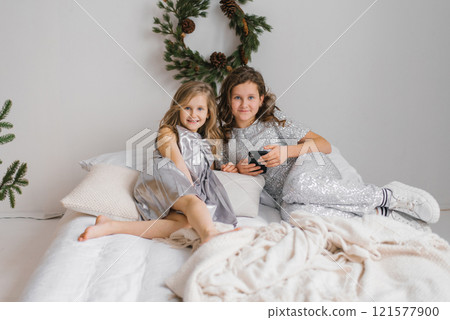Two young sisters girls are sitting on bed, one of them holding smartphone. The room is decorated with Christmas trees and pine cones, giving it a festive atmosphere 121577900