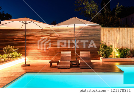 Elegant poolside area with lounge chairs and umbrellas at night. Modern outdoor design illuminated with warm lighting 121578551
