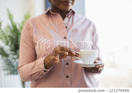 African American woman in office environment holding coffee cup with relaxed, contemplative expression, suggesting business break or leisure moment. Emphasizes calm, reflective office atmosphere. 121579569