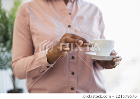 African American woman in office environment holding coffee cup with relaxed, contemplative expression, suggesting business break or leisure moment. Emphasizes calm, reflective office atmosphere. 121579570