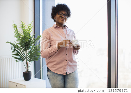 African American woman wearing glasses standing by window holding coffee cup, expressing confidence and calm in office setting. Indoor plant adds touch of nature. 121579571