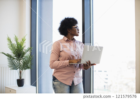 Young African American businesswoman holding laptop standing by office window. Casual attire, focused expression, natural light. Indoor plant adds greenery to modern office setting. Young African American businesswoman holding laptop standing by office window. Casual attire, focused expression, natural light. Indoor plant adds greenery to modern office setting. 121579576