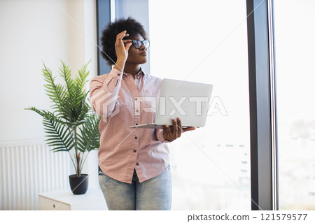 African American businesswoman standing near window holding laptop, wearing casual attire. Daylight and indoor plant create modern, productive atmosphere. 121579577