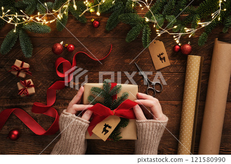 Preparing for the holiday, gift wrapping. Women's hands tie a bow on a gift box. Table with handmade items, rolls of kraft brown paper, red velvet ribbon, labels, scissors. 121580990