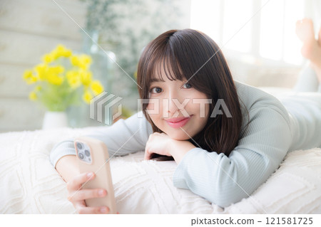 Close-up of a young woman lying in bed, holding a smartphone and looking at the camera with a smile - Spring image Close-up of a young woman lying in bed, holding a smartphone and looking at the camera with a smile - Spring image 121581725