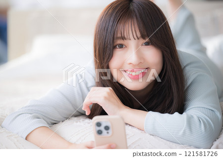Close-up of a young woman lying in bed, holding a smartphone and looking at the camera with a smile Close-up of a young woman lying in bed, holding a smartphone and looking at the camera with a smile 121581726