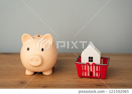 Piggy bank with house in shopping cart. Concept of buying, selling or renting a home. Mortgage, loan, real estate Piggy bank with house in shopping cart. Concept of buying, selling or renting a home. Mortgage, loan, real estate 121582065